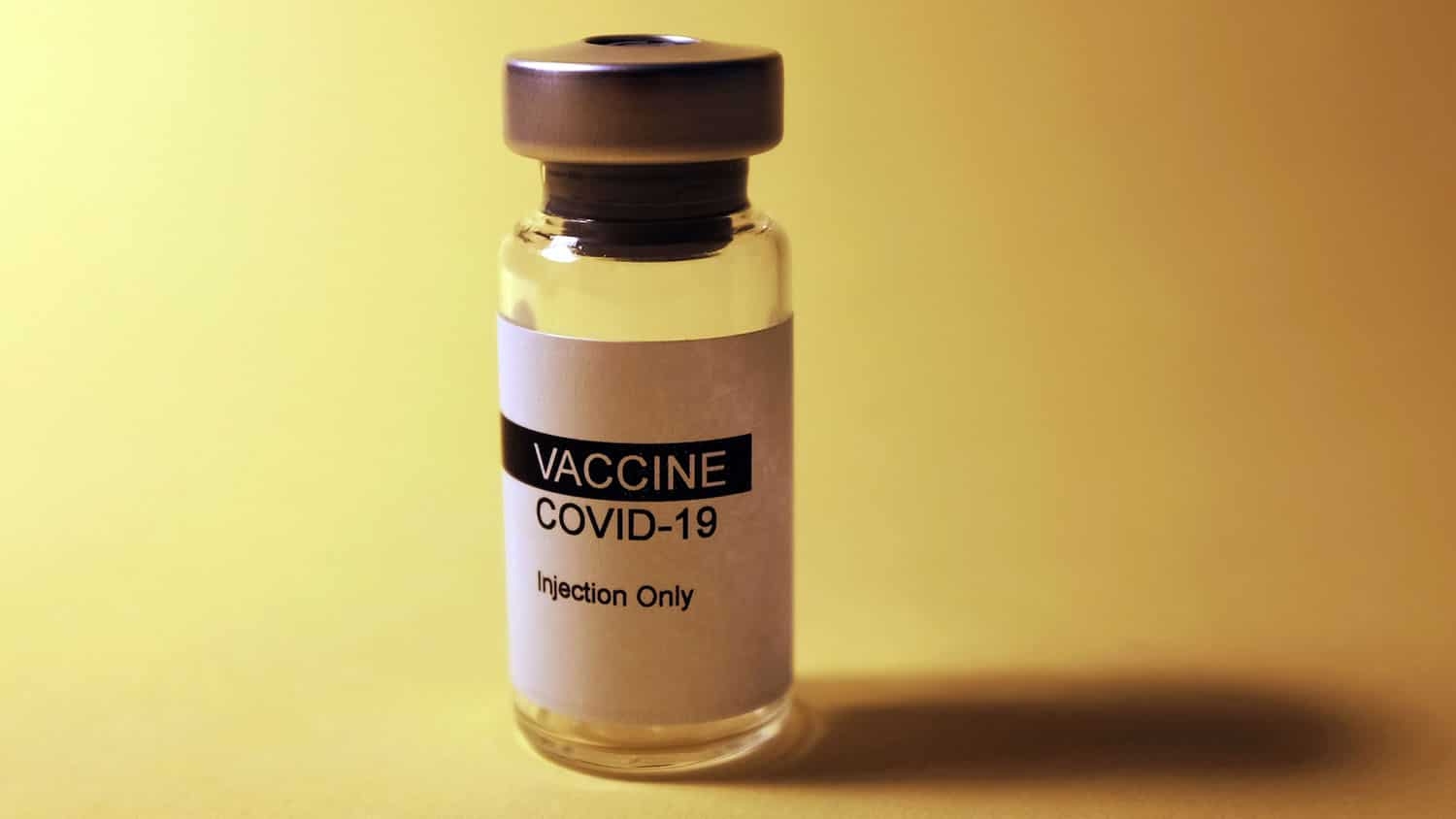 a vial labelled "COVID-19 Vaccine" sits on a table against a yellow backdrop